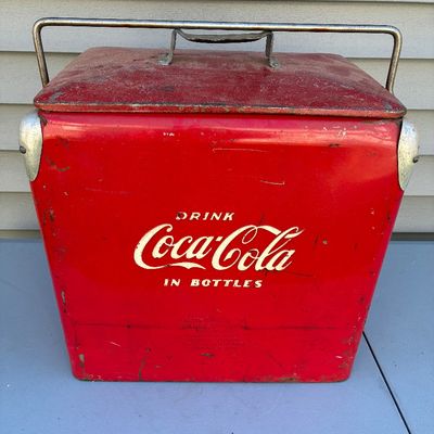 Sale Photo Thumbnail #123: Vintage red metal Coca-Cola portable ice cooler with metal carry handle, featuring classic "Drink Coca-Cola In Bottles" embossed lettering and age-appropriate paint wear and patina.