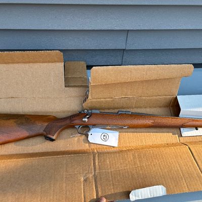 Sale Photo Thumbnail #59: Bolt-action rifle featuring a checkered wooden stock and rubber butt pad, displayed inside a cardboard box with an identification tag.