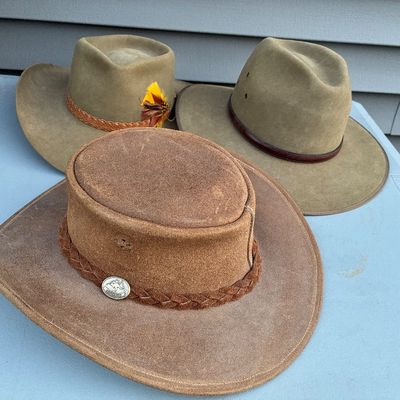 Sale Photo Thumbnail #114: Trio of vintage leather and felt Australian outback-style hats, including an Akubra-style hat with a feather embellishment and a suede hat with a braided band.