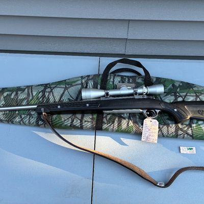 Sale Photo Thumbnail #61: Ruger bolt-action rifle featuring a synthetic stock, stainless steel barrel, attached scope, and a leather sling, displayed on top of a camouflage soft gun case.