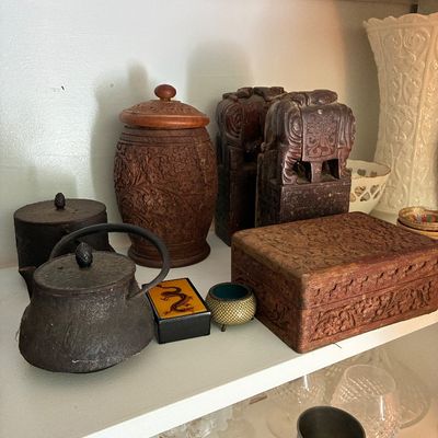 Sale Photo Thumbnail #38: Collection of Asian and global decorative items, including hand-carved wooden boxes, carved elephant bookends, cast iron teapots, and a small cloisonné bowl displayed on a shelf.