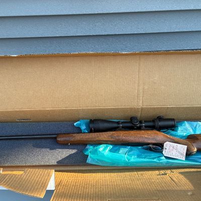 Sale Photo Thumbnail #57: Bolt-action hunting rifle with a wood stock and mounted scope, presented inside its foam-lined cardboard storage box.