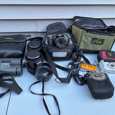 Sale Photo Thumbnail #68: Collection of photography gear and optics, including a Nikon Coolpix DSLR camera, point-and-shoot digital cameras, extra lenses, L.L. Bean binoculars, and several padded carrying bags.