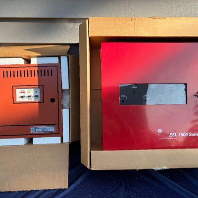 Sale Photo Thumbnail #24: Two vintage red fire alarm control panels, including a Fire Lite Alarms MP-12 module and a GE ESL 1500 Series panel, both in original cardboard boxes with packaging.