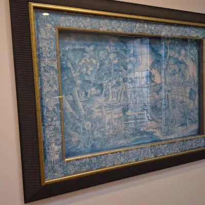 Sale Photo Thumbnail #26: Framed blue and white toile fabric art depicting a forest scene with animals and a fountain, set in a textured dark frame with gold trim.