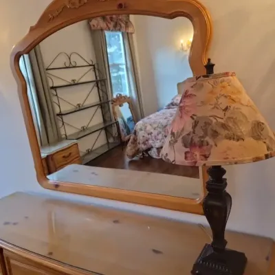 Sale Photo Thumbnail #44: Traditional knotty pine dresser with an attached arched mirror, topped with a protective glass surface and paired with a dark metal lamp featuring a floral shade.