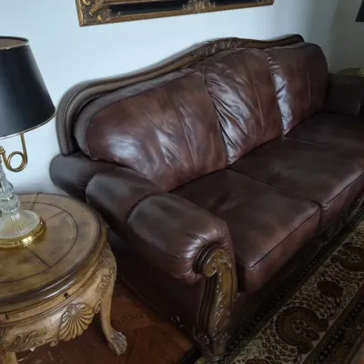 Sale Photo Thumbnail #17: Ornate dark brown leather sofa featuring carved wood trim and scrolled details, flanked by a matching round side table with a black and brass lamp.