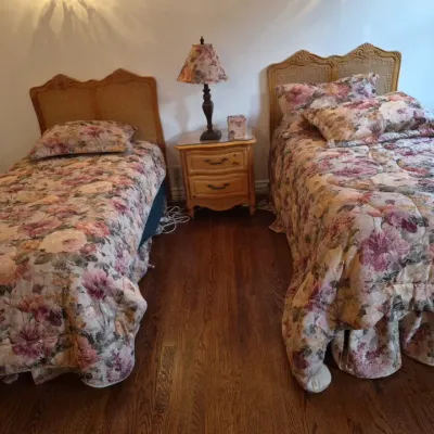 Sale Photo Thumbnail #46: Pair of French Provincial style twin bed frames with cane headboards, dressed in floral comforters, flank a matching two-drawer nightstand with a table lamp.