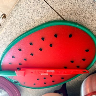 Novelty watermelon-shaped cutting board with a green rim, black seed details, and a matching red utility knife in a protective sheath.