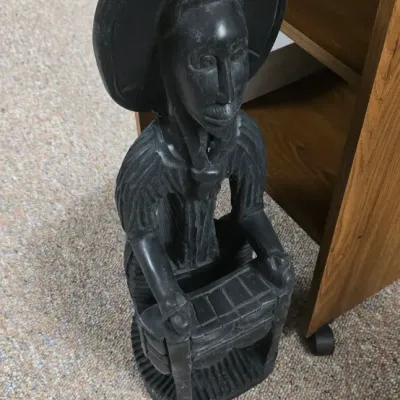 Sale Photo Thumbnail #37: Hand-carved dark wood African-style figure depicting a musician playing an instrument, standing beside a wooden shelf.