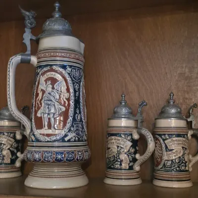 Sale Photo Thumbnail #19: Collection of German beer steins featuring a large central stein flanked by six smaller matching steins, all with detailed pewter lids and reliefs.