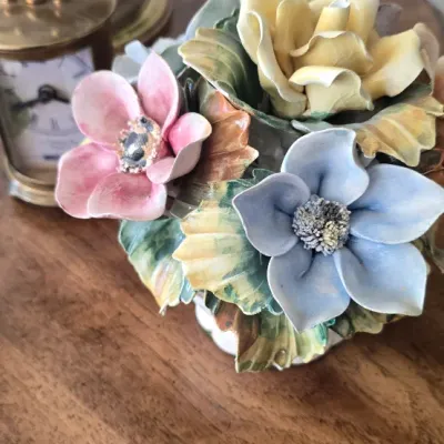 Sale Photo Thumbnail #59: Hand-painted Capodimonte style porcelain floral bouquet sculpture featuring colorful, delicate blooms and green leaves, displayed on a wooden surface alongside a vintage table clock and lamp base.