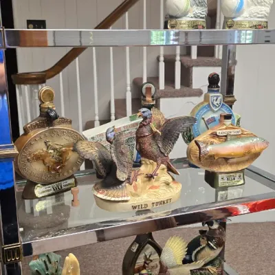 Sale Photo Thumbnail #34: Collection of vintage collectible liquor decanters, featuring various animals and wildlife motifs including poodles, wild turkeys, and trout, displayed on a glass shelving unit.