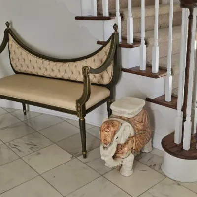 Sale Photo Thumbnail #2: Neoclassical style settee bench with button-tufted backrest upholstery and a carved ceramic elephant garden stool beside a staircase.