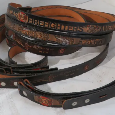 Collection of embossed leather firefighter-themed belts, featuring detailed tooling of fire trucks, Maltese crosses, and text reading...
