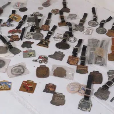 Large collection of vintage belt buckles, watch fobs, and pins featuring trucks, heavy machinery, and Mack bulldog themes.