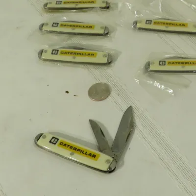 Collection of vintage Caterpillar advertising pocket knives with cream-colored synthetic handles, mostly in original plastic sleeves.