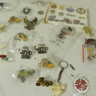 Collection of vintage novelty enamel lapel pins, keychains, and badges featuring heavy machinery, vehicles, Harley-Davidson, and fire...