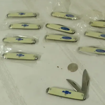 Collection of small pocket knives featuring Chevrolet and Ford logos on cream-colored handles, mostly sealed in plastic packaging.