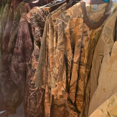 Sale Photo Thumbnail #86: Collection of camouflage hunting clothing hanging on a clothing rack, including jackets, t-shirts, and coveralls in various realtree and woodland patterns.