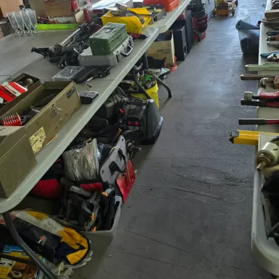 Sale Photo Thumbnail #62: Garage filled with various tools, hardware, toolboxes, and equipment laid out on long folding tables and stored underneath.