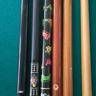 Sale Photo Thumbnail #113: Collection of assorted billiard cues resting on green felt, featuring wooden shafts and novelty designs including a Budweiser beer themed cue stick.