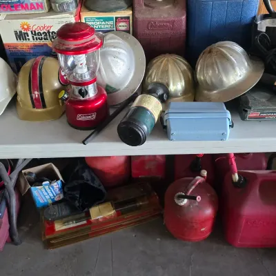 Sale Photo Thumbnail #56: Collection of vintage outdoor gear and hard hats displayed on folding tables, including a red Coleman lantern, metal construction helmets, and propane canisters.