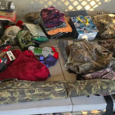 Sale Photo Thumbnail #97: Assortment of outdoor gear, including camouflage apparel, a soft gun case, a black motorcycle helmet, and various folded clothing items displayed on a table.