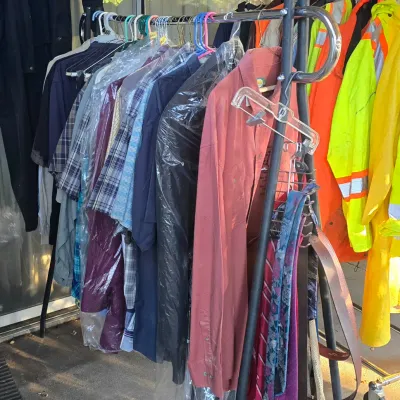 Sale Photo Thumbnail #101: A metal rolling clothing rack filled with men's apparel, including button-up shirts, dry-cleaned garments, ties, belts, and high-visibility work jackets.