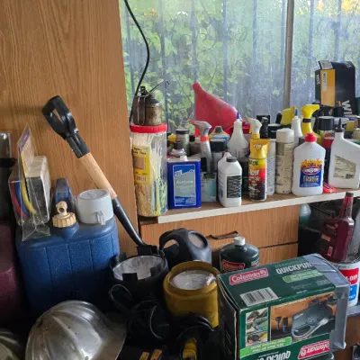 Sale Photo Thumbnail #58: Assortment of workshop and outdoor supplies, including a Coleman propane stove, QuickPump, metal hard hat, gas cans, and various chemicals.