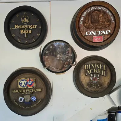 Sale Photo Thumbnail #105: Collection of five vintage wall signs and clocks, featuring German brewery barrel-head signs including Henninger Bräu and Hacker-Pschorr alongside a Taylor deer thermometer.