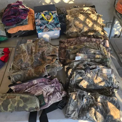 Sale Photo Thumbnail #98: Large assortment of outdoor and hunting apparel, featuring camouflage pants, jackets, ghillie suit material, waist packs, and folded flannel shirts.