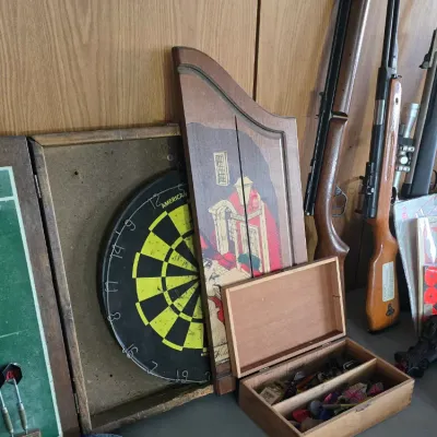 Sale Photo Thumbnail #60: Vintage dartboard cabinet set with a small wooden dart storage box, pitched horseshoe set, and several air rifles leaned against the wall.
