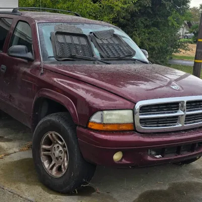 Sale Photo Thumbnail #117: Maroon late 1990s to early 2000s Dodge Durango SUV parked on a concrete driveway, featuring a chrome front grille, roof rails, and rubber floor mats propped on the windshield.