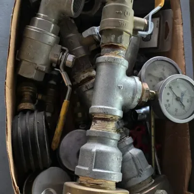 Sale Photo Thumbnail #18: Cardboard box filled with assorted metal plumbing fittings, pipe valves, unions, and pressure gauges showing signs of prior use.