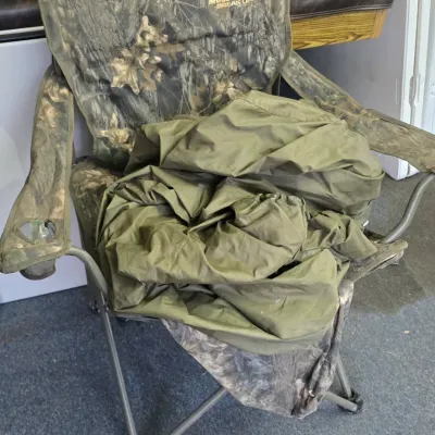 Sale Photo Thumbnail #39: Mossy Oak Break-Up camouflage folding camp chair with armrest cup holders and an olive green tarp or cover placed on top.