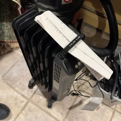 Black digital oil-filled radiator space heater with paper instructions tucked under the handle, sitting next to a Craftsman wet/dry vacuum.