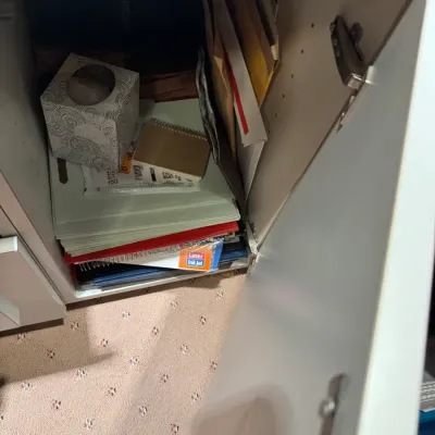 Open white storage cabinet filled with office supplies, including folders, notebooks, envelopes, and a tissue box cover.