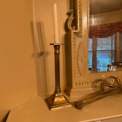 Neoclassical style brass column candlestick holder with a beaded square base, set on a fireplace mantel next to a vintage carved mirror.