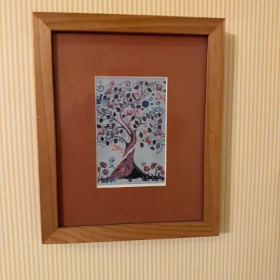 Framed folk art print depicting a stylized tree of life with birds and flowers, set within a wide reddish-brown mat and light wood frame.