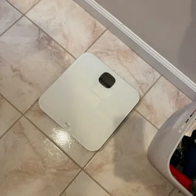 White Fitbit smart bathroom scale sitting on a tiled floor.