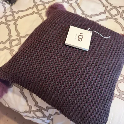UGG Home knit throw pillow in deep purple featuring fuzzy pom-pom corners and original retail tag attached.