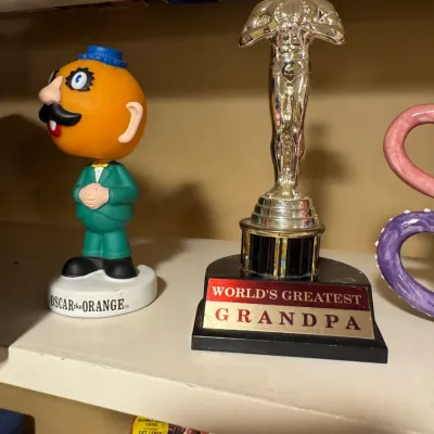 A vintage Oscar the Orange bobblehead paired with a gold-tone 'World's Greatest Grandpa' Oscar-style trophy statue on a...