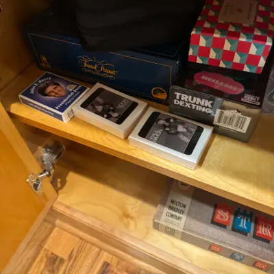 Collection of tabletop games and playing cards on a cabinet shelf, including Trivial Pursuit, Trunk Texting, and novelty card decks.