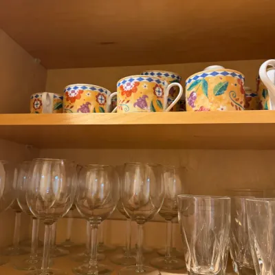 Collection of glassware and ceramic tableware displayed on wooden cabinet shelves, including wine glasses, tumblers, and colorful...