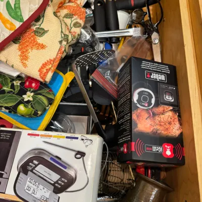 Kitchen drawer filled with assorted utensils and gadgets, including a boxed Weber iGrill mini thermometer, Sharper Image digital cooking...