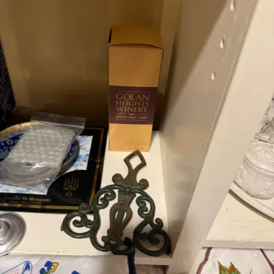 Golan Heights Winery boxed bottle alongside cast iron trivet, Judaica plates, and household items on a shelf.