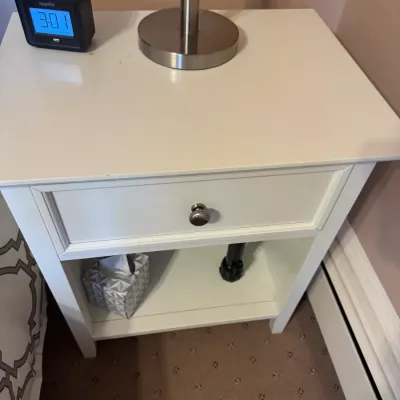 White single-drawer nightstand with brushed metal hardware and lower open shelf, holding a small digital alarm clock and table lamp base.
