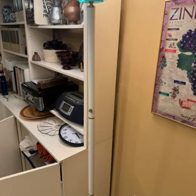 Modern white torchiere floor lamp standing beside a white shelving unit filled with household items and electronics.
