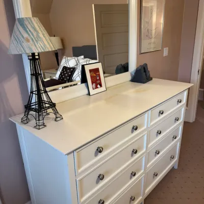 White wooden six-drawer dresser with an attached rectangular mirror and silver knobs. A novelty wire Eiffel Tower table lamp and small...